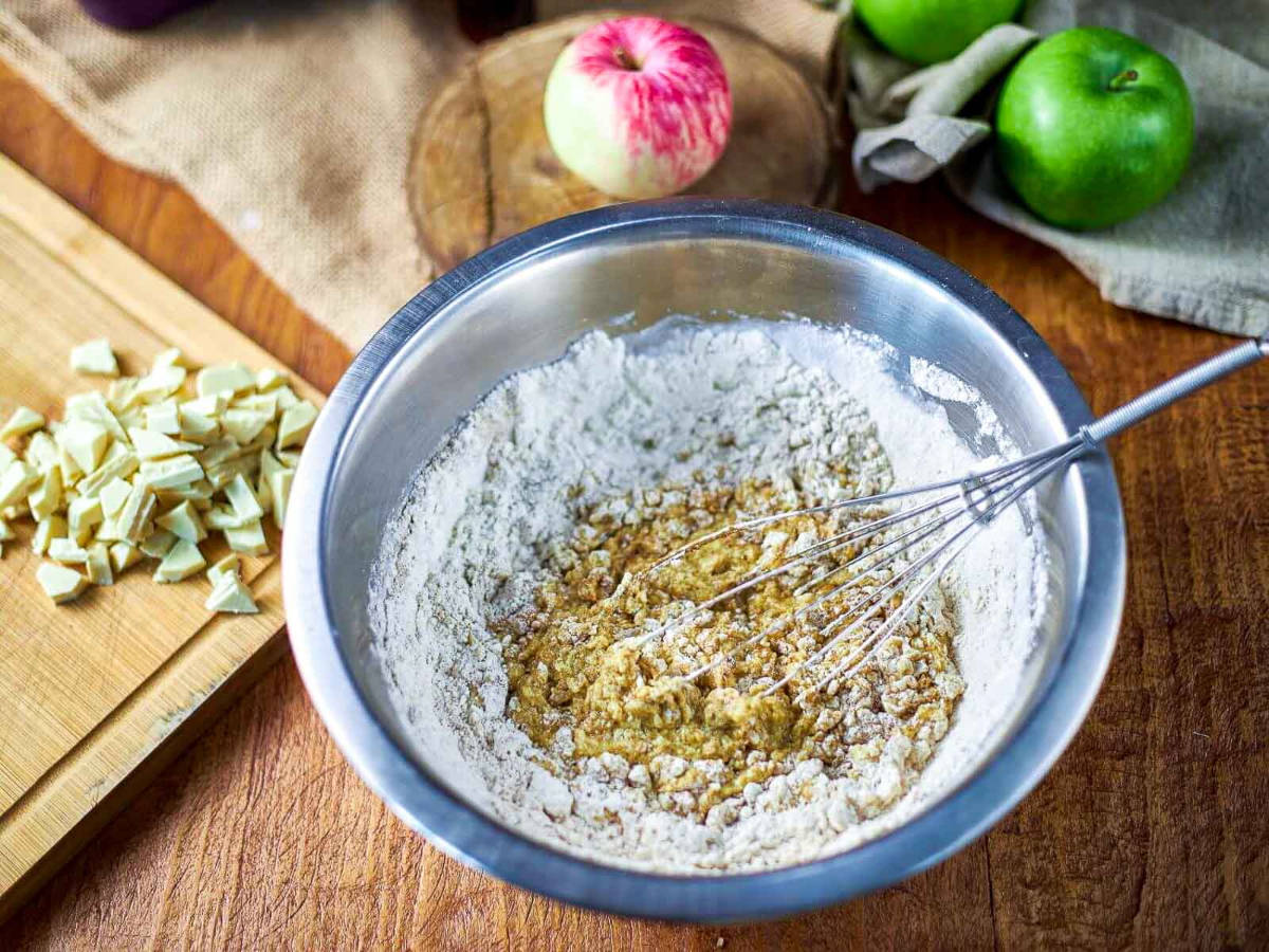 A whisk folding flour into a batter in a metal bowl, with chopped white chocolate on a cutting board to the side.