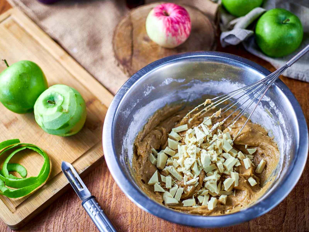 A whisk folding chopped white chocolate into blondie batter in a metal bowl, next to peeled green apples and a vegetable peeler on a cutting board.