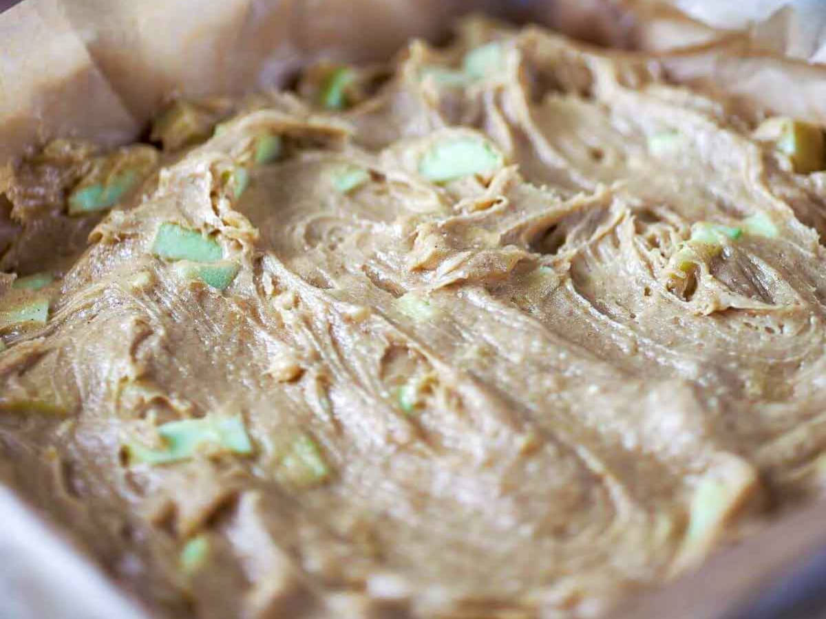 Blondie batter spread into a parchment-lined baking pan, with chunks of green apple visible throughout.