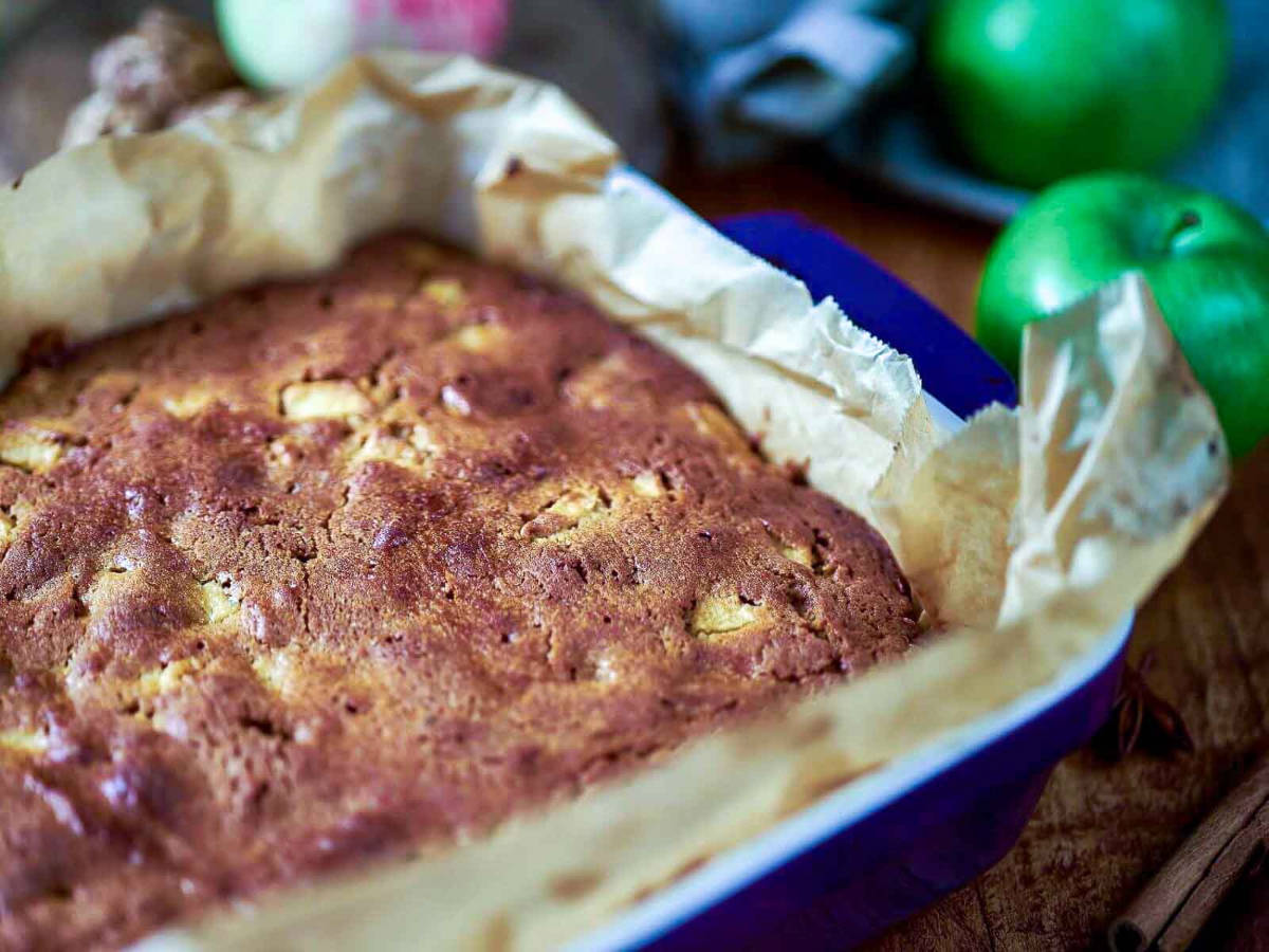 A pan of freshly baked blondies with a golden brown top, lined with parchment paper, next to green apples.