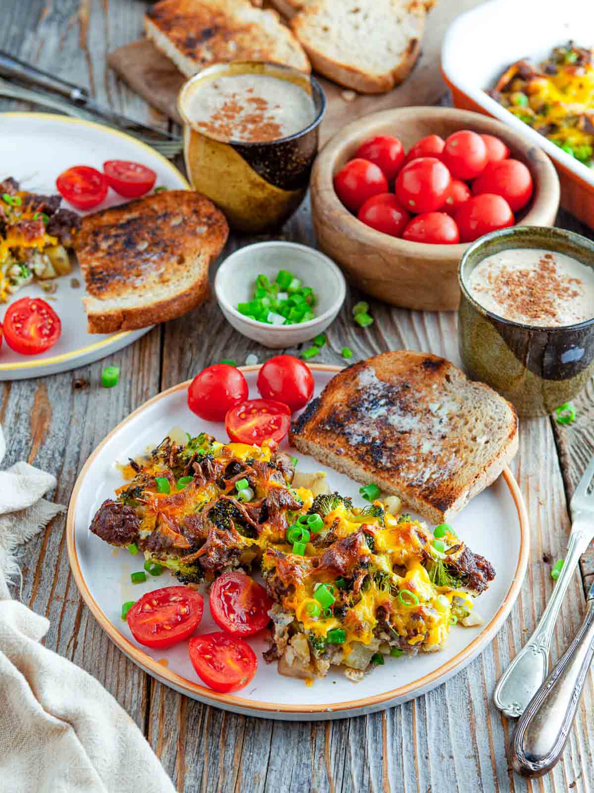 A plate of potato sausage and broccoli breakfast casserole next to toasted sourdough and halved cherry tomatoes, with a mug of coffee and more casserole in the background