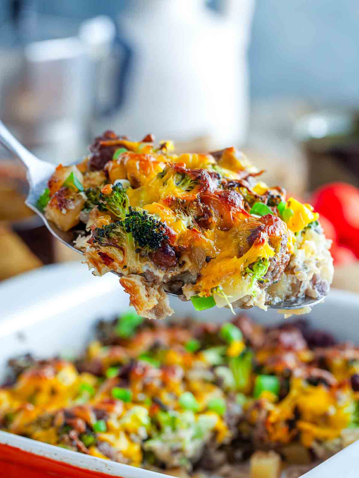 A spoon lifting a scoop of potato sausage broccoli breakfast casserole out of the baking dish.