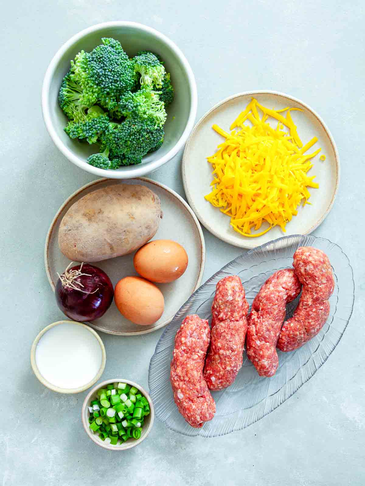 Ingredients for potato sausage broccoli breakfast casserole laid out on a light gray surface: broccoli, shredded cheddar, a russet potato, eggs, a red onion, milk, chopped chives, and raw Italian sausage links.