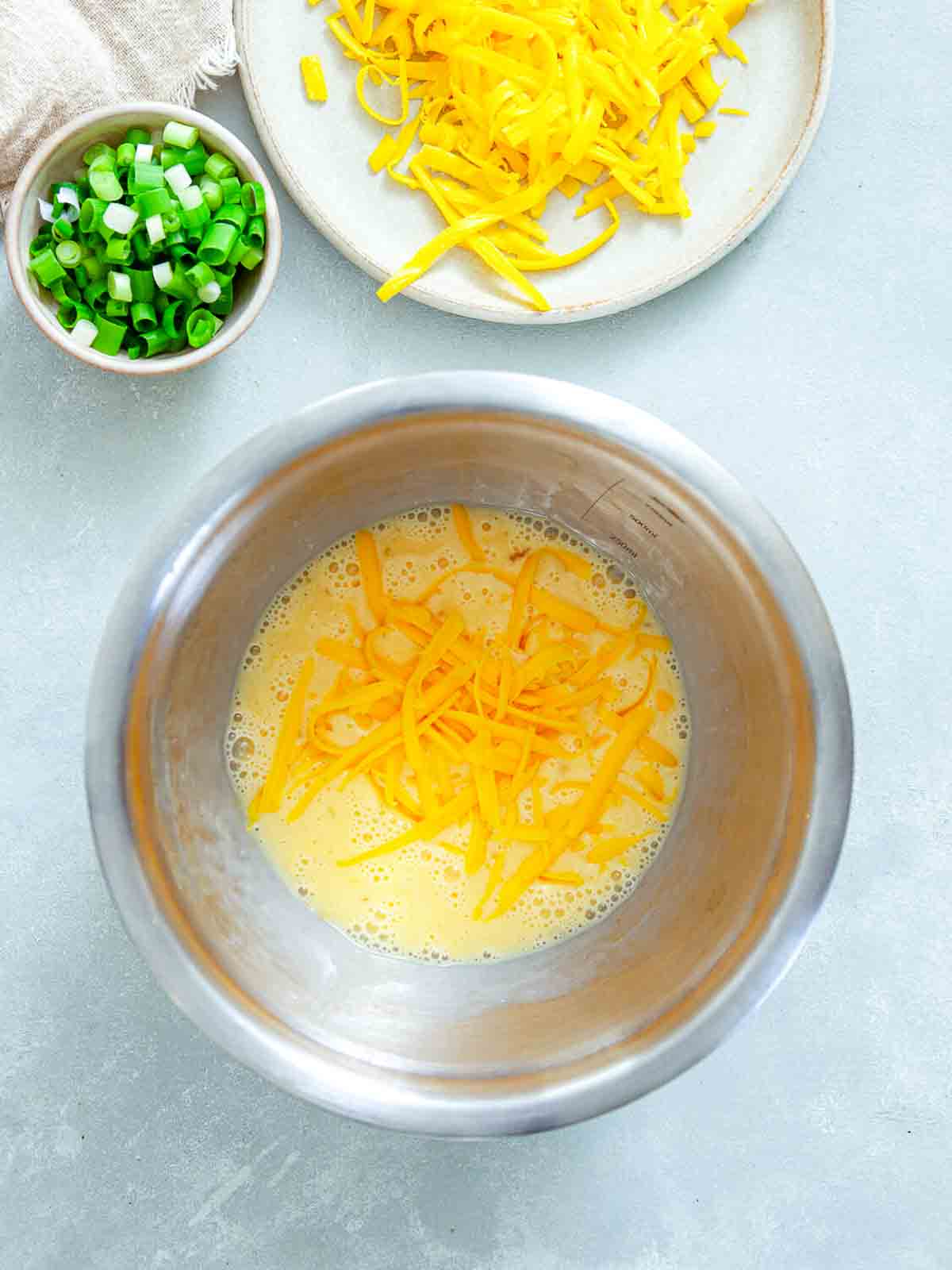 Shredded cheddar cheese added to beaten eggs in a mixing bowl, with chopped green onions nearby.
