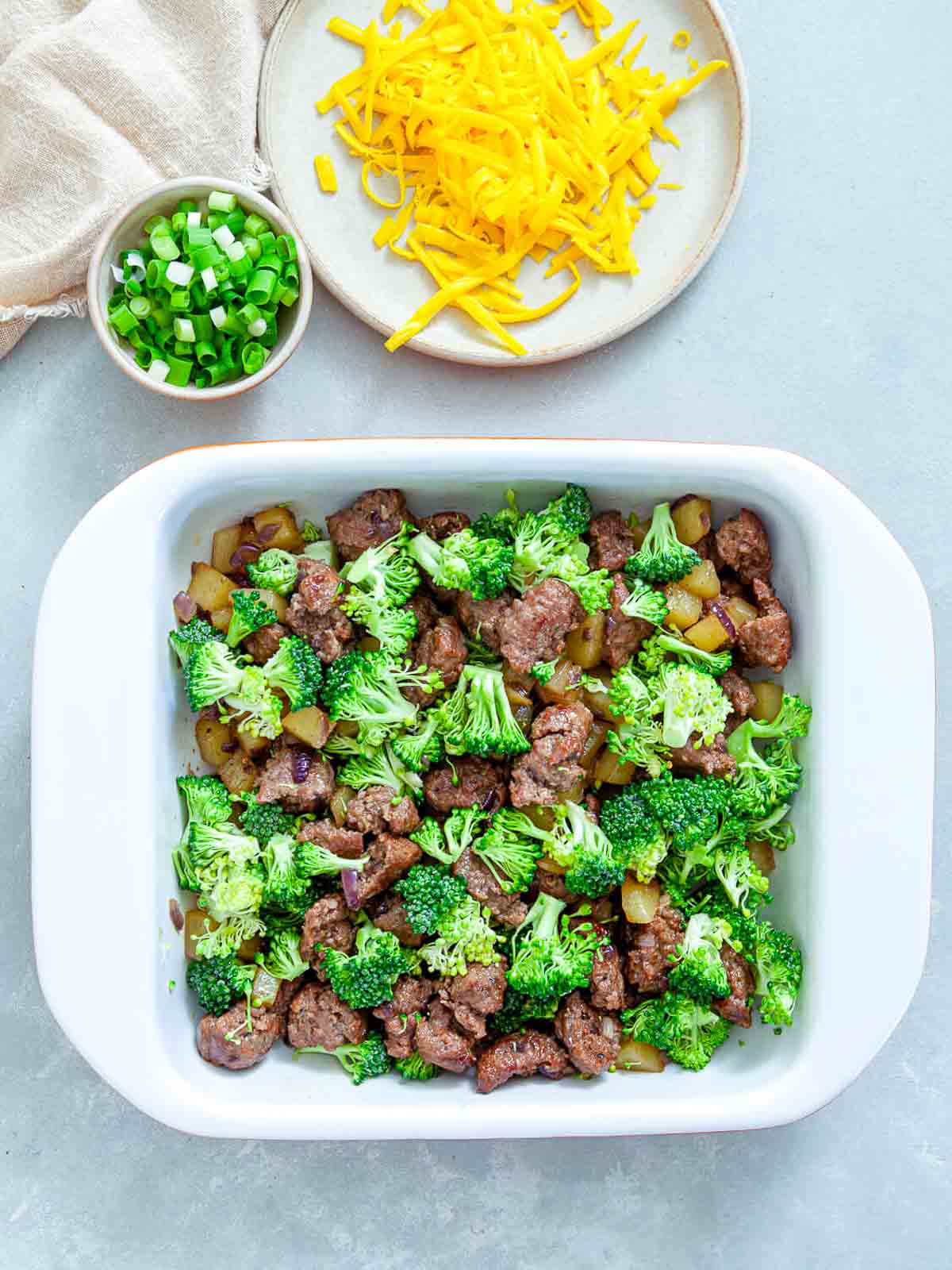 Sausage, potato, and broccoli assembled in a white baking dish before the egg mixture and cheese are added.