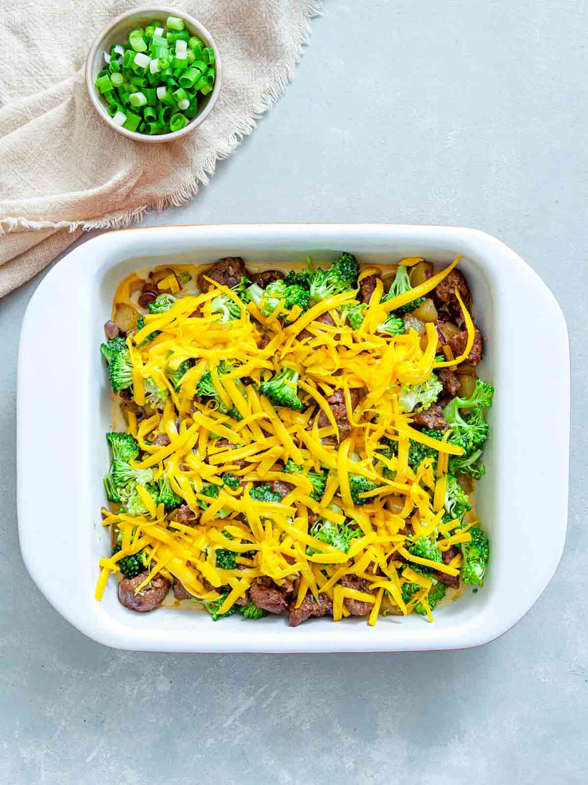 Potato sausage broccoli breakfast casserole topped with shredded cheddar cheese in a white baking dish before baking.