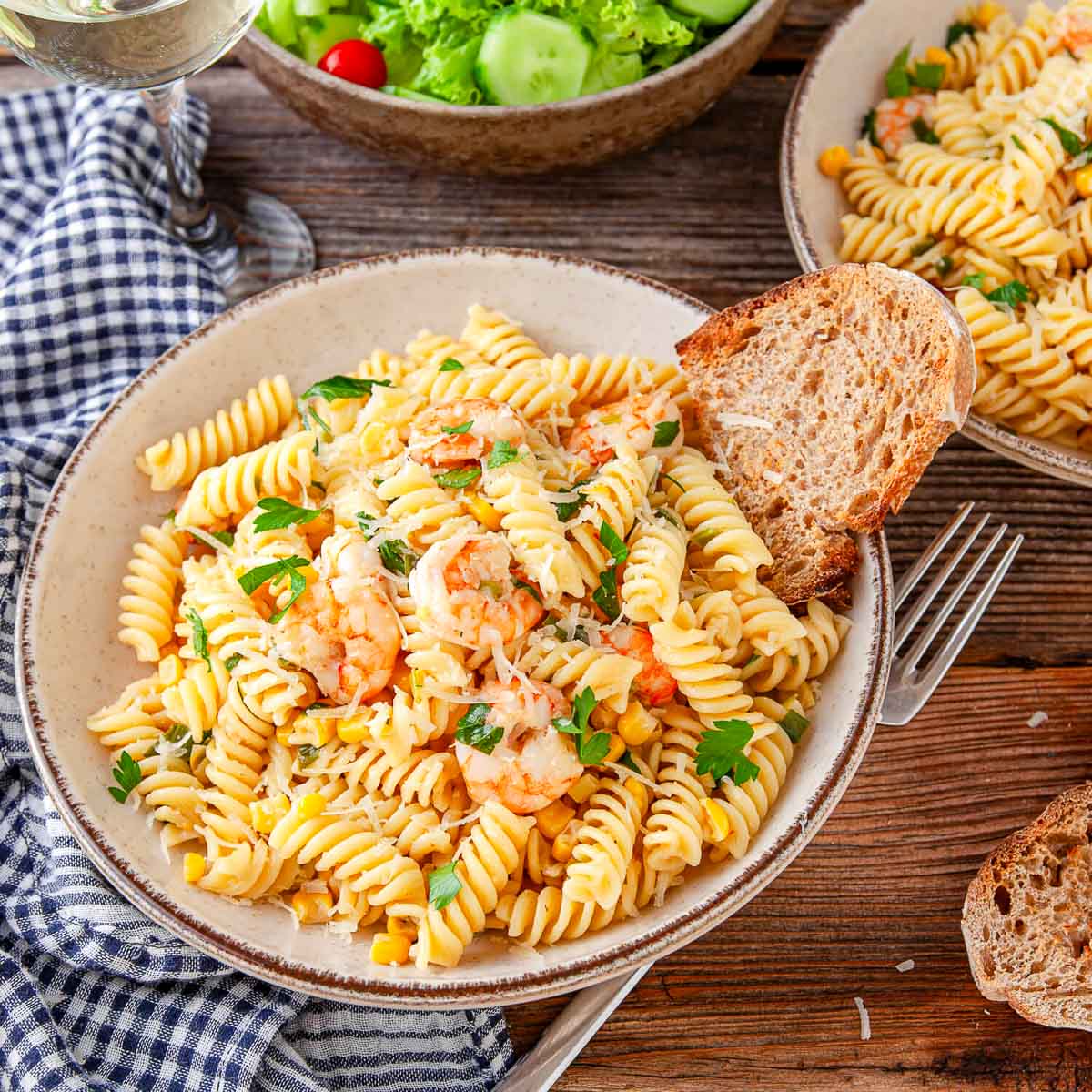 Overhead view of a bowl of shrimp and corn rotini topped with parmesan and parsley, served with a slice of crusty bread and a side salad.