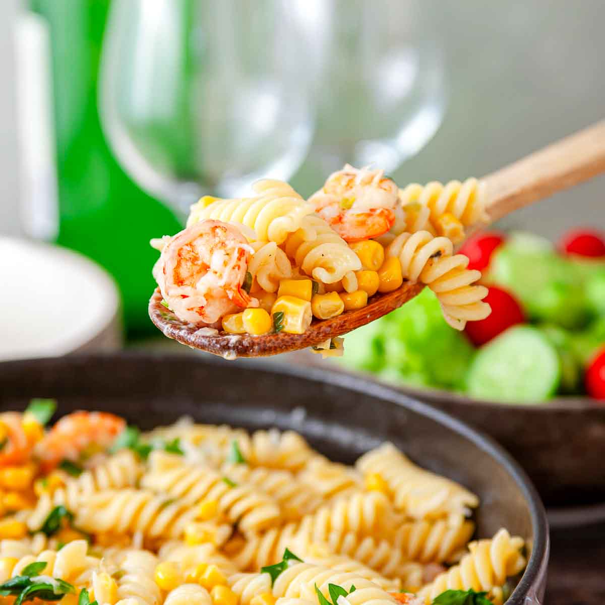 A wooden spoon holding a bite of pasta with shrimp and corn kernels above the finished dish.