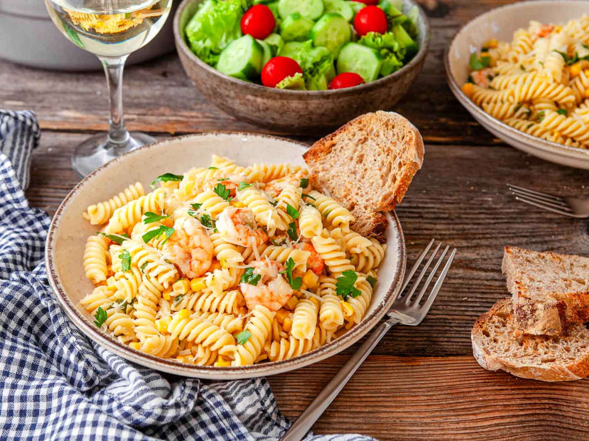 Bowl of rotini pasta with shrimp, corn, and fresh parsley on a wood table with bread and a cucumber tomato salad.
