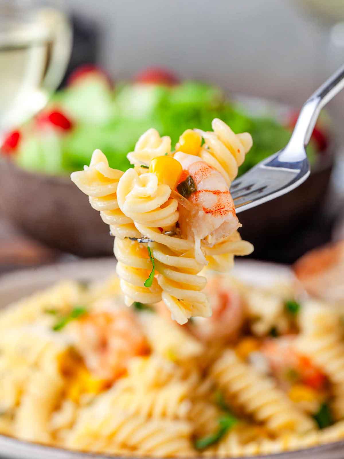 A forkful of rotini pasta with a shrimp and a piece of corn lifted from the bowl.
