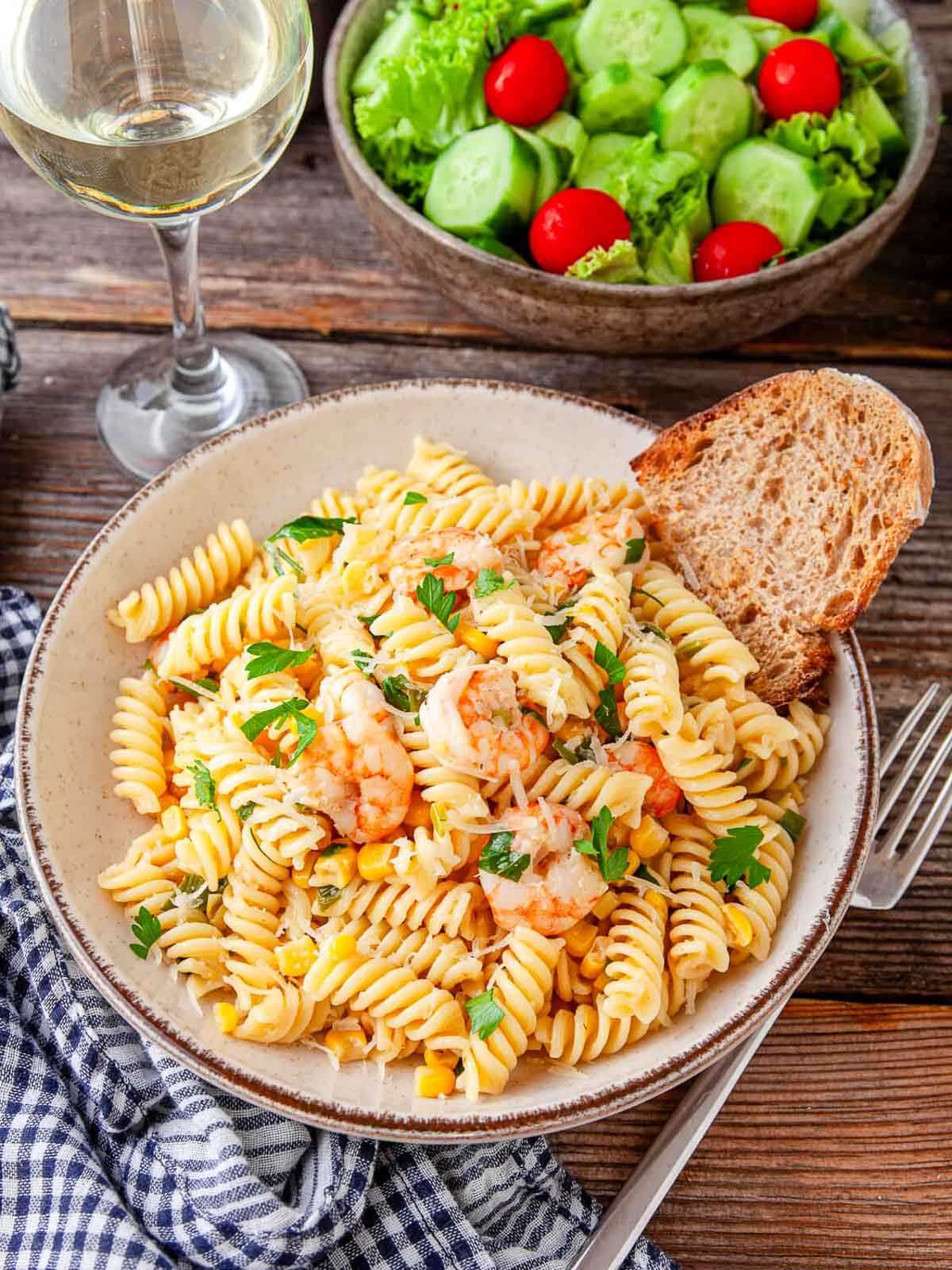 Side view of a bowl of pasta with shrimp, corn, and parmesan next to a glass of white wine and a green salad.