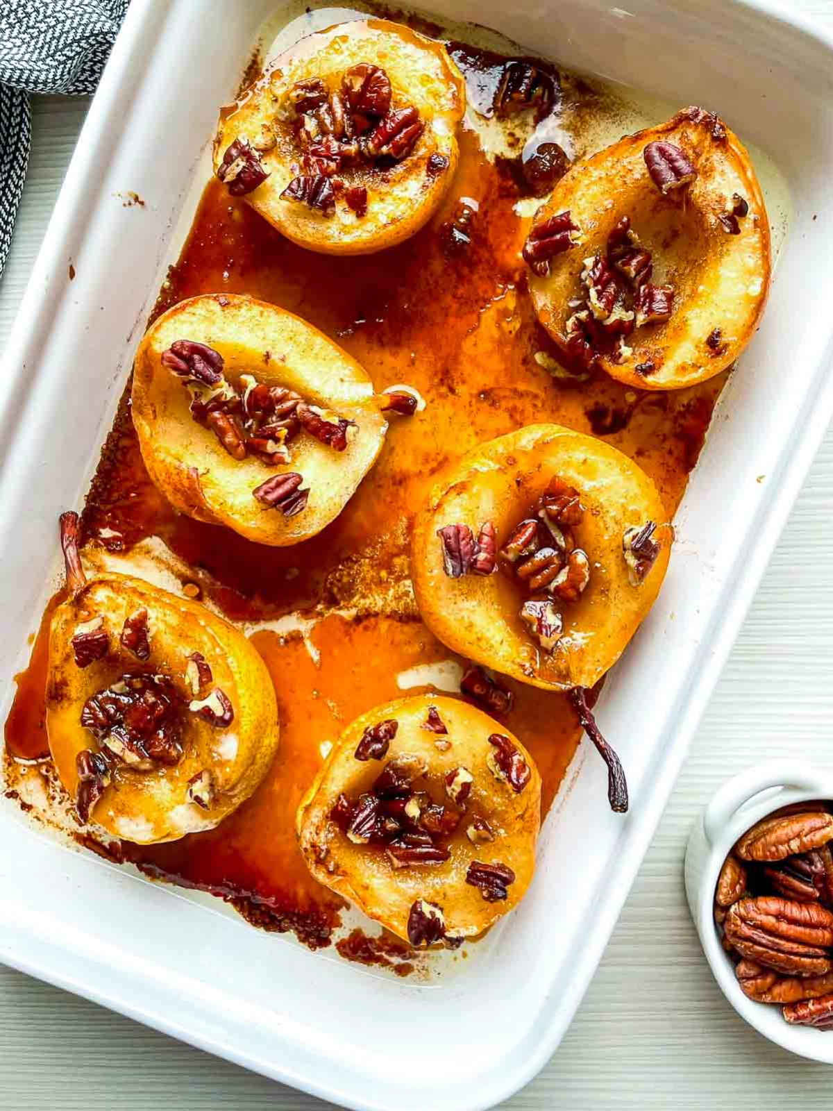Six caramelized pear halves topped with chopped pecans in a white baking dish, surrounded by a pool of brown sugar syrup.