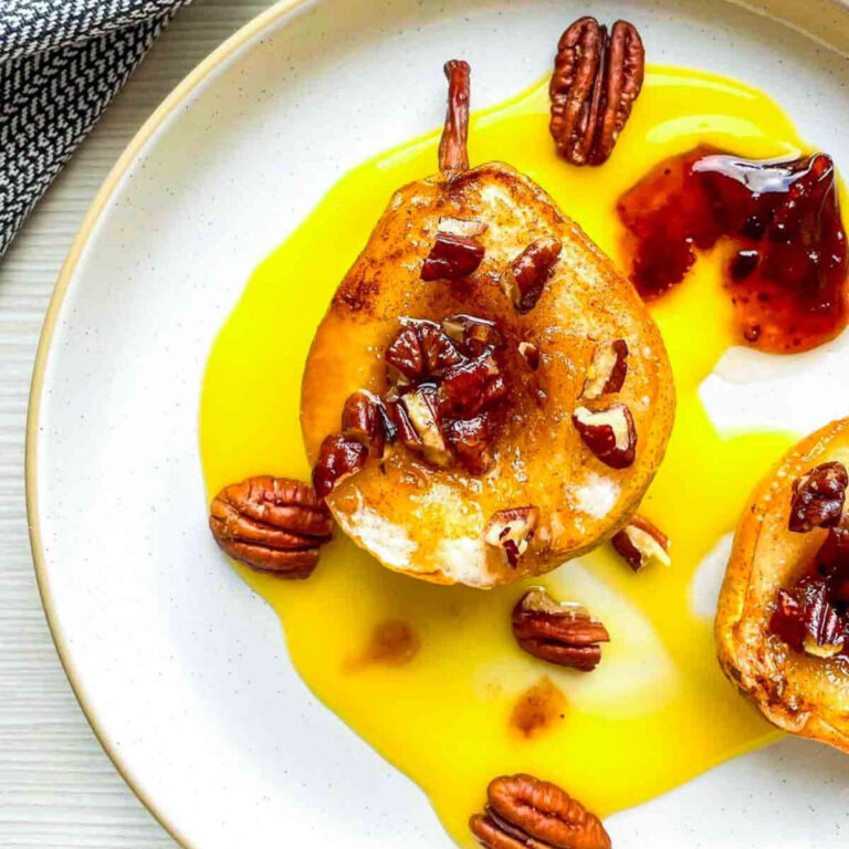 A plated serving of two baked pear halves with pecans in a pool of syrup, next to a small dab of red preserves, with the baking dish visible in the background.