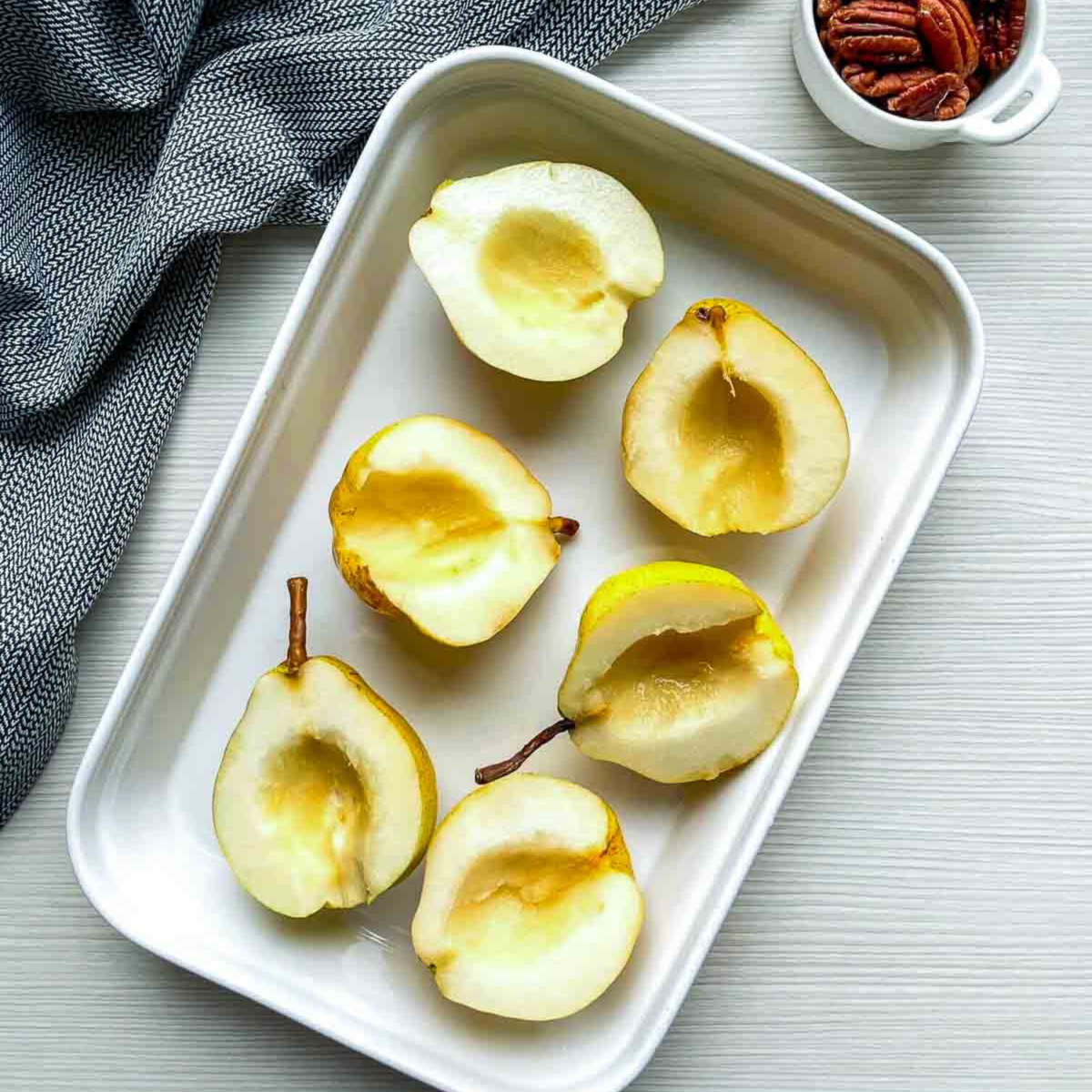 Six pear halves with the cores scooped out, arranged in a white baking dish before being tossed with the coating.