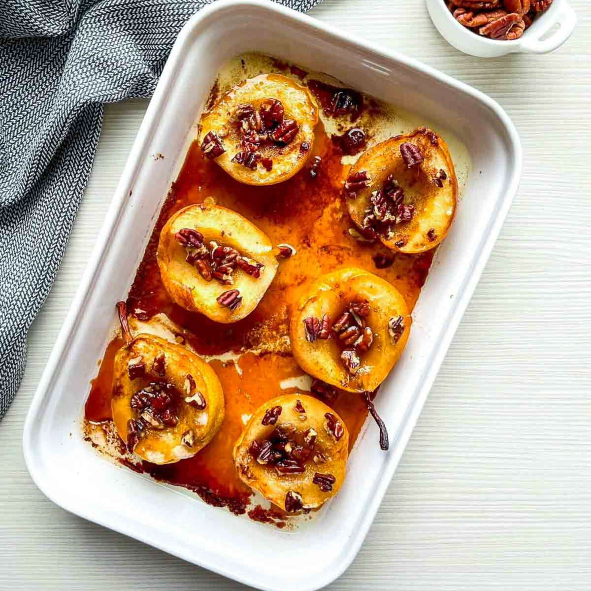 Golden, caramelized pear halves topped with pecans, fresh out of the oven in a white baking dish with syrup pooling around them.
