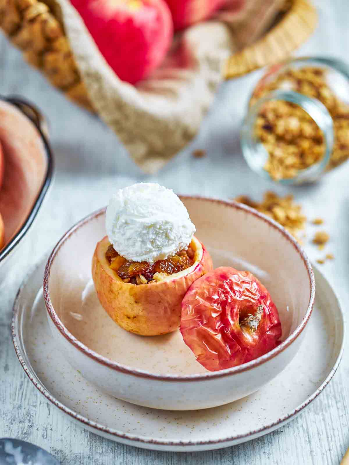  a single granola stuffed baked apple topped with a scoop of vanilla ice cream, in a bowl next to a second baked apple