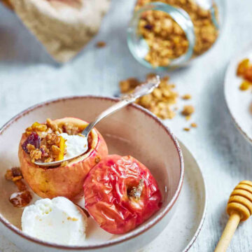 a spoon lifting granola filling from a baked apple, with a scoop of ice cream and a plate of dried fruit nearby
