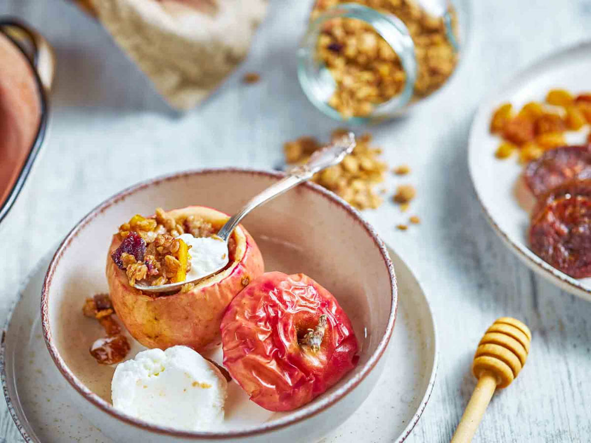 a spoon lifting granola filling from a baked apple, with a scoop of ice cream and a plate of dried fruit nearby