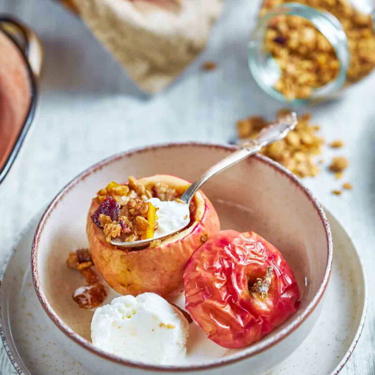 a spoon lifting granola filling from a baked apple topped with crème fraîche, with a small plate of dried fruit alongside