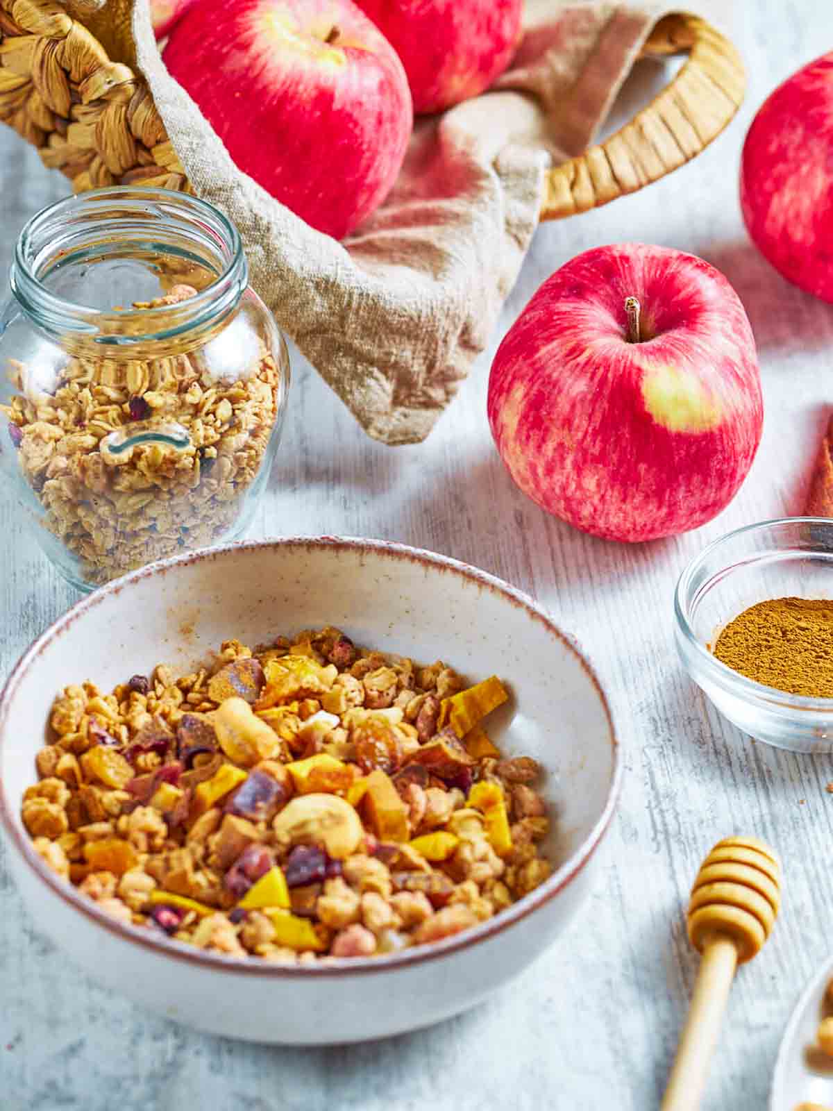granola, raisins, dried fruit, and cinnamon mixed together in a white bowl, with apples in a basket in the background