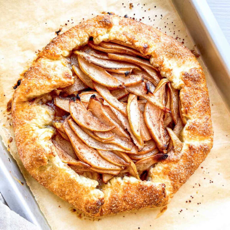 A whole rustic pear galette with a golden, sugar-crusted edge and thinly sliced spiced pears fanned across the center, on a parchment-lined baking sheet.