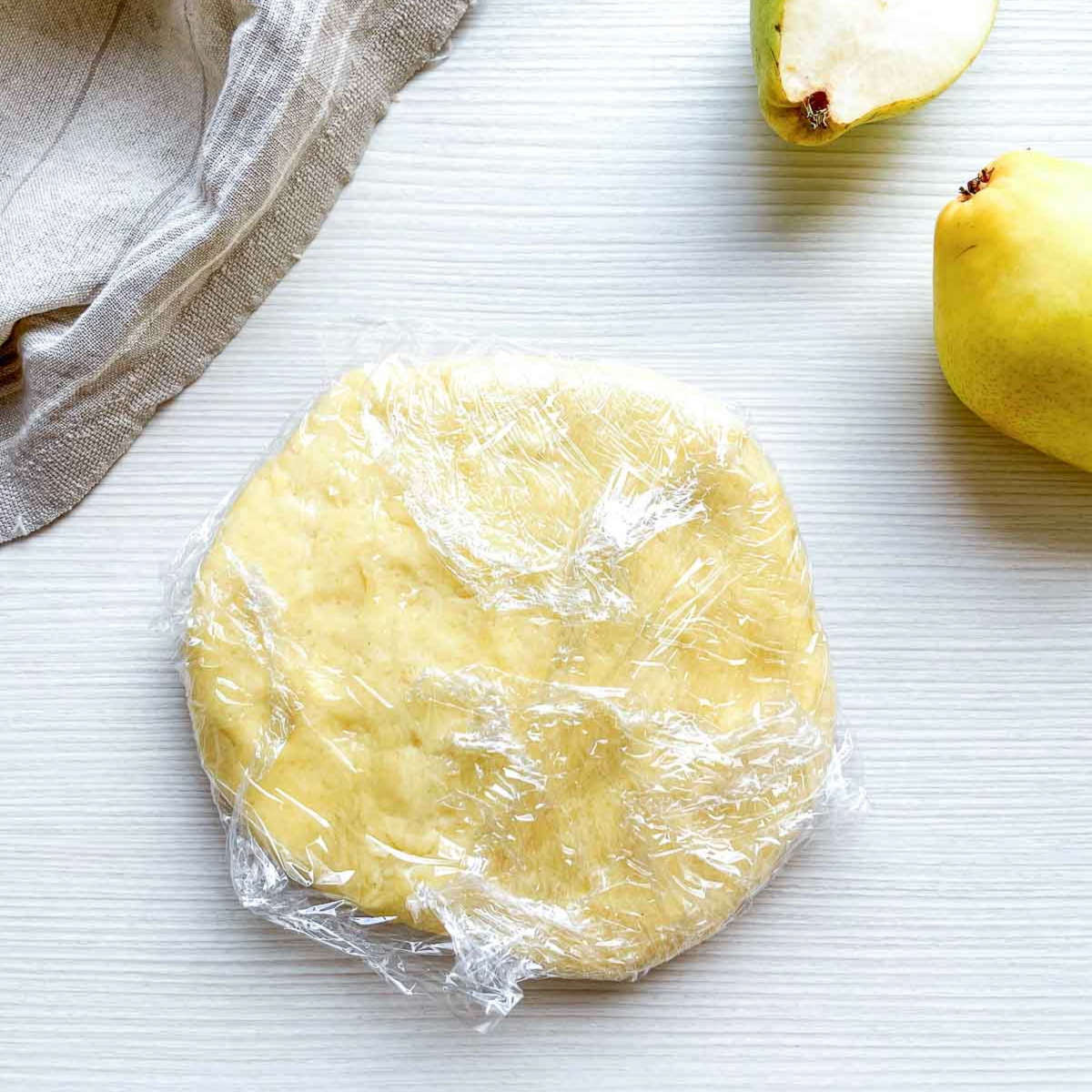 A flattened disc of pie dough wrapped tightly in plastic wrap, resting on a white countertop before chilling.