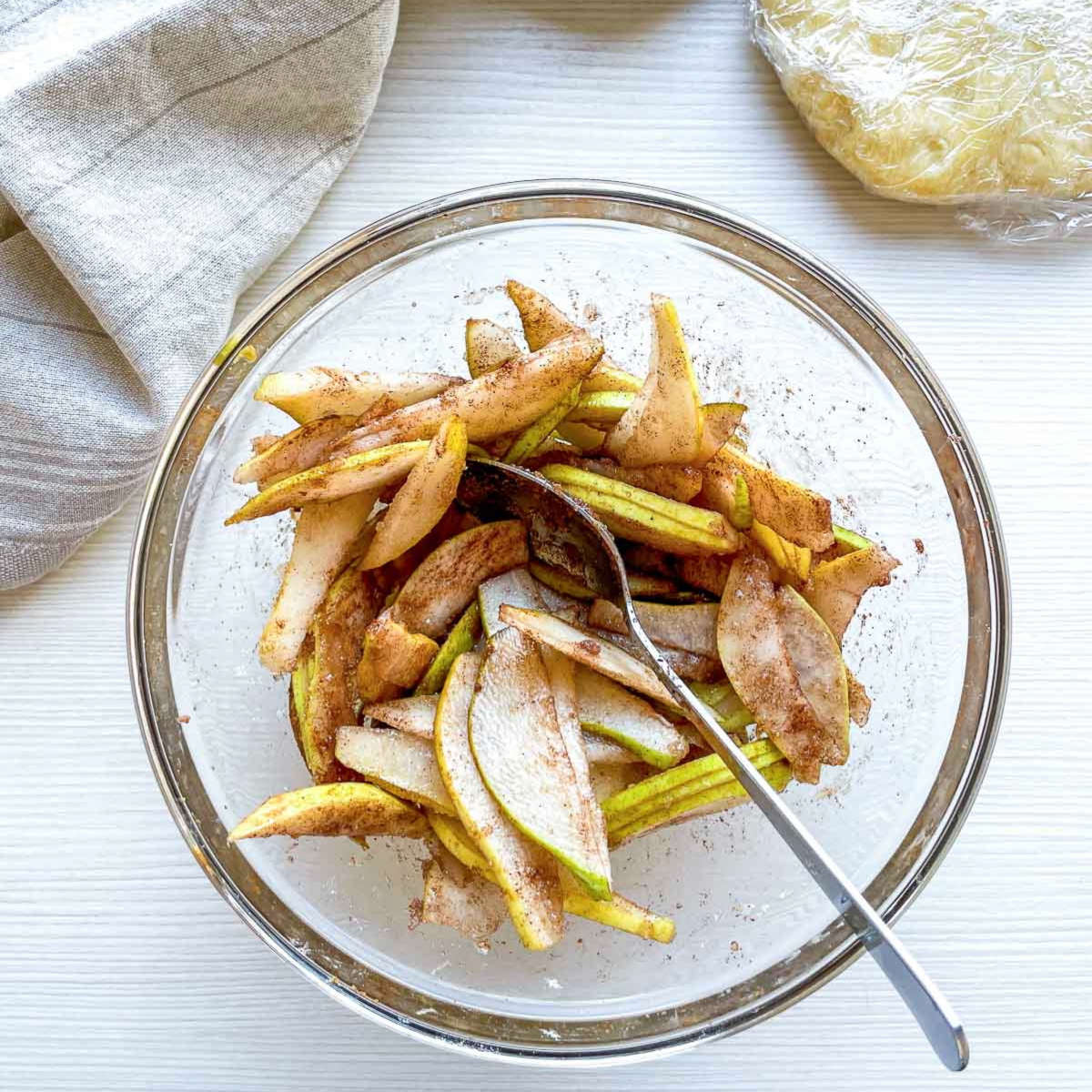 Thin pear slices coated in cinnamon and sugar, tossed together in a glass bowl with a spoon.