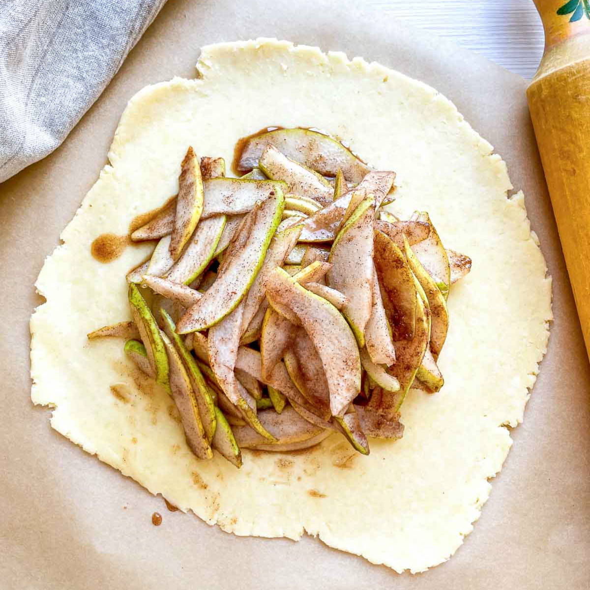 Cinnamon-spiced pear slices piled in the center of a rolled-out circle of pie dough on parchment paper.