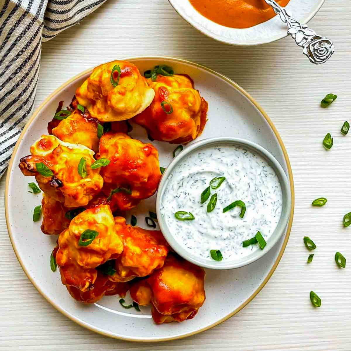 Buffalo cauliflower wings on plate with a bowl of creamy herb dip and scattered green onions