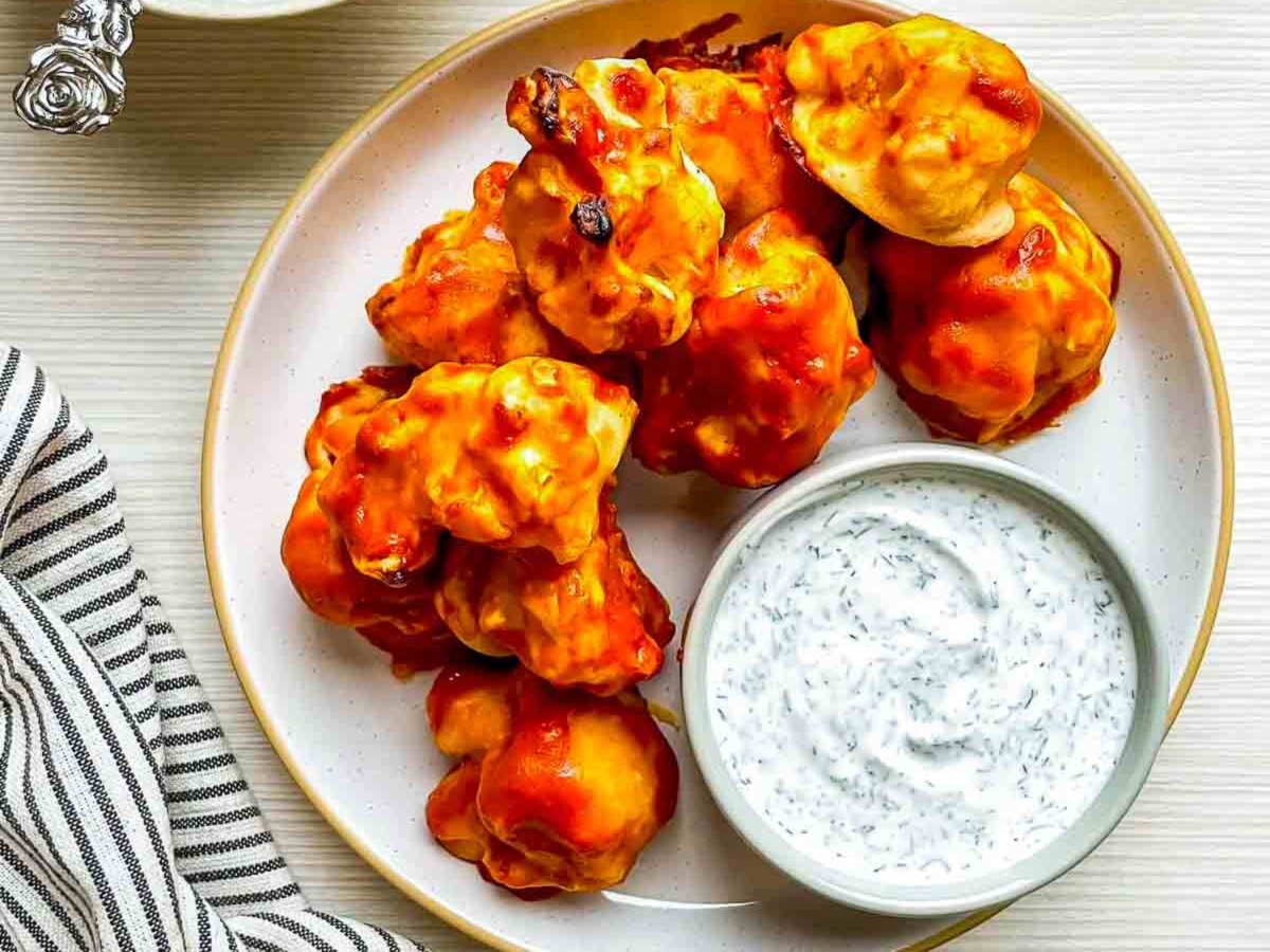 Overhead view of golden orange cauliflower wings beside a bowl of herb dip on a speckled plate