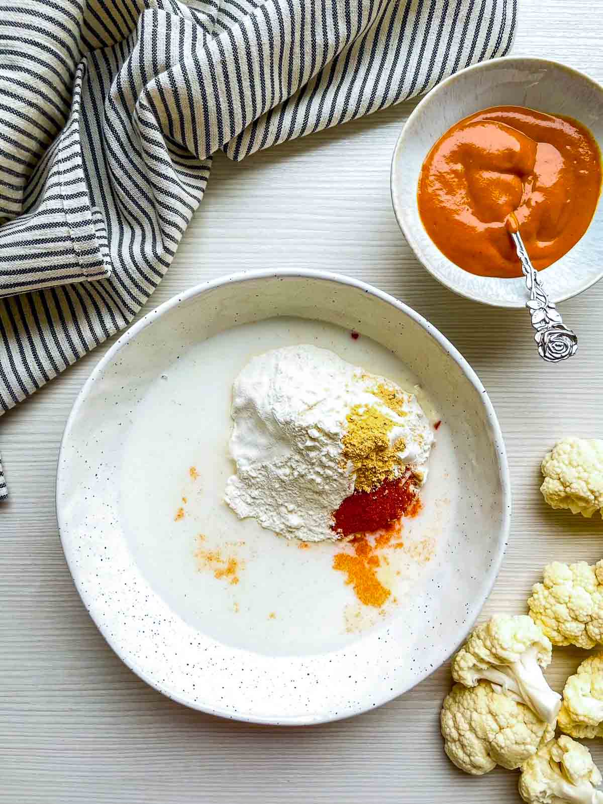 Speckled bowl of milk with flour, paprika, and garlic powder piled on top, with raw florets and a bowl of buffalo sauce nearby