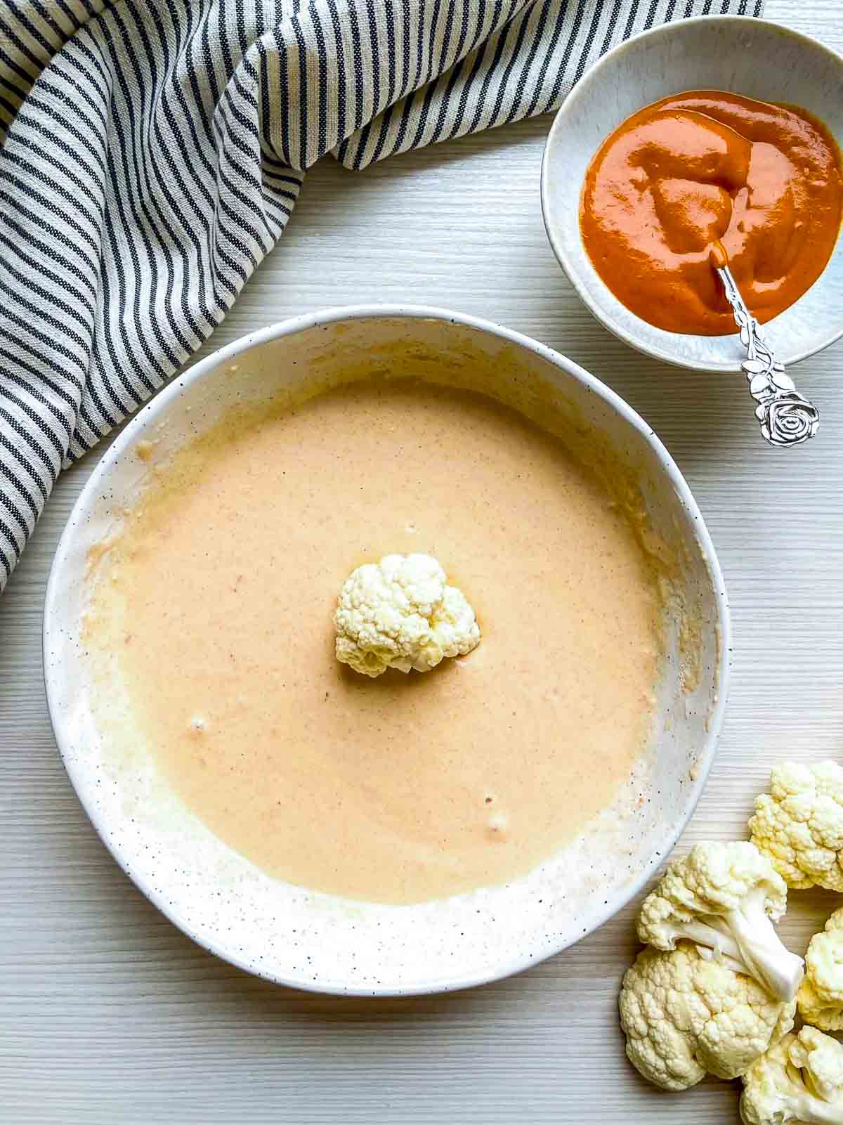 Smooth pale orange batter in a speckled bowl with a single raw cauliflower floret resting in the center