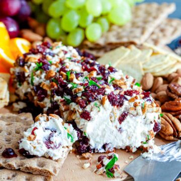 Orange cranberry cheese ball coated in chopped pecans, dried cranberries, and parsley on a wooden board with crispbread, grapes, and orange slices.