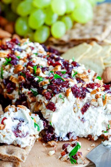 Orange cranberry cheese ball coated in chopped pecans, dried cranberries, and parsley on a wooden board with crispbread, grapes, and orange slices.