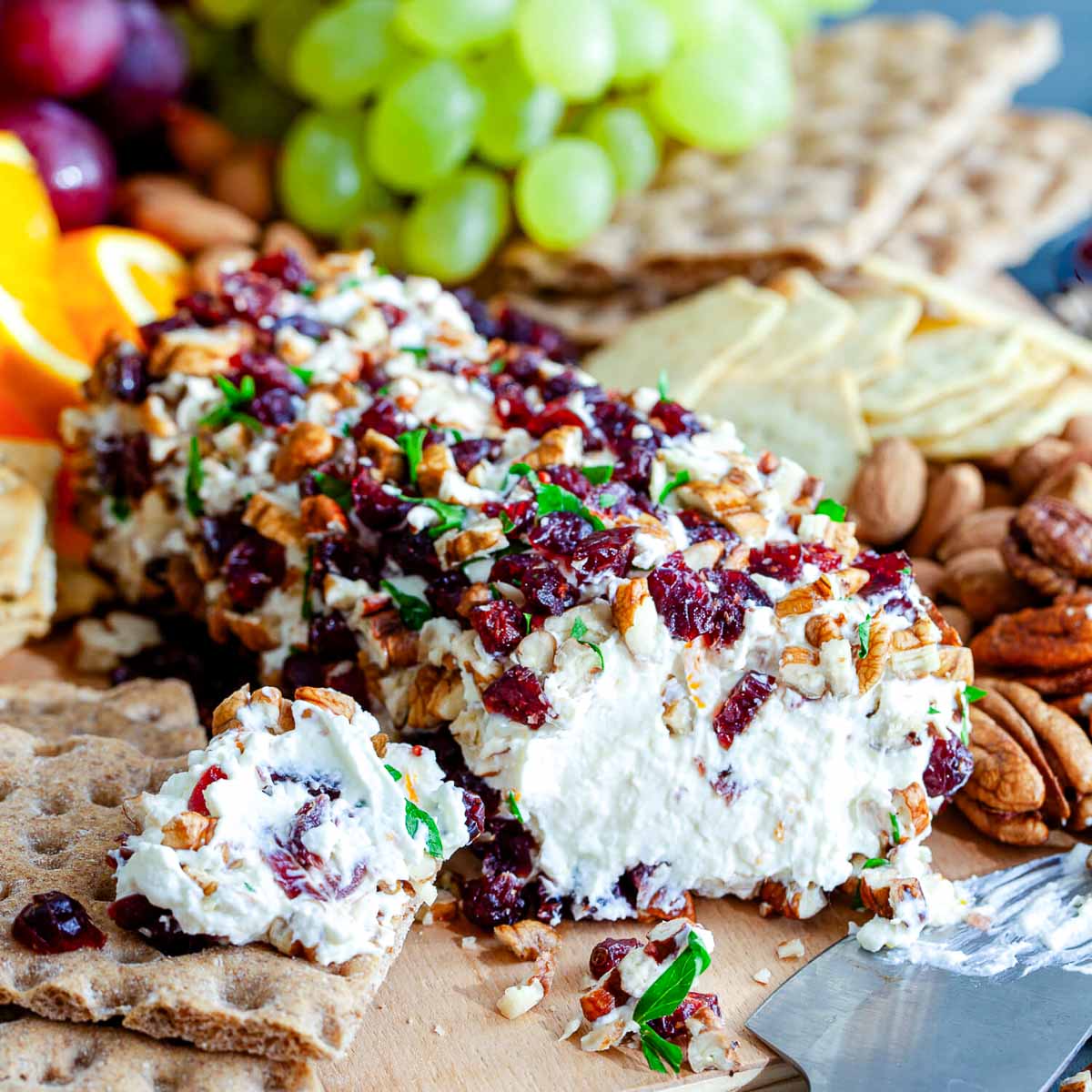 Orange cranberry cheese ball coated in chopped pecans, dried cranberries, and parsley on a wooden board with crispbread, grapes, and orange slices.