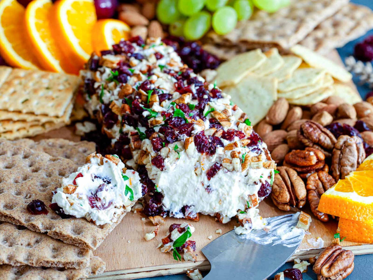 A cut cheese log showing a creamy white center, crusted with pecans and cranberries, surrounded by crackers, almonds, pecans, and orange slices.