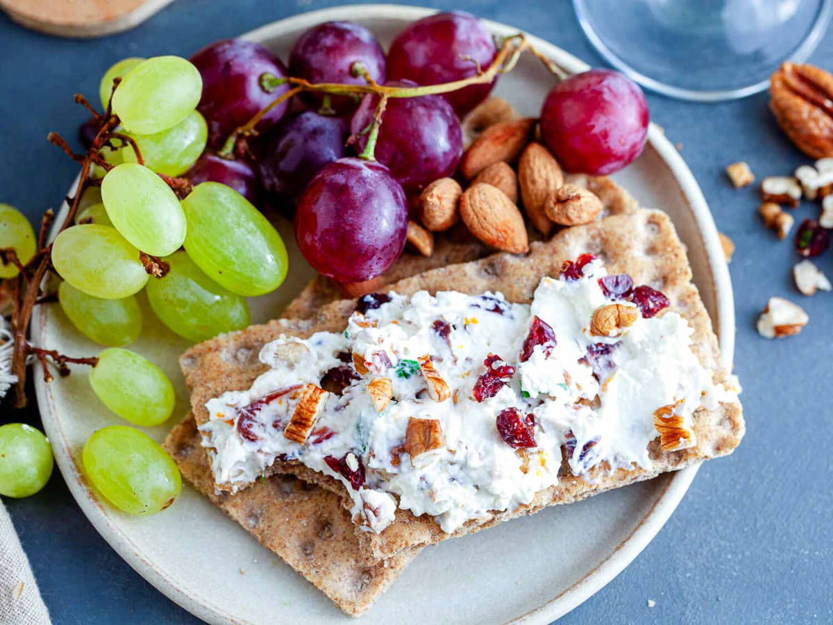 Creamy cheese spread studded with cranberries and pecans on rye crispbread, served on a plate with red and green grapes and almonds.