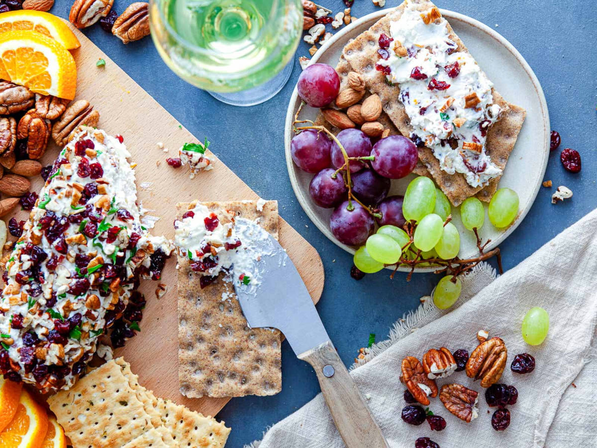 Overhead view of a pecan and cranberry crusted cheese log on a wooden board next to a plate of grapes, almonds, and crispbread with spread, a glass of white wine, and a linen napkin.