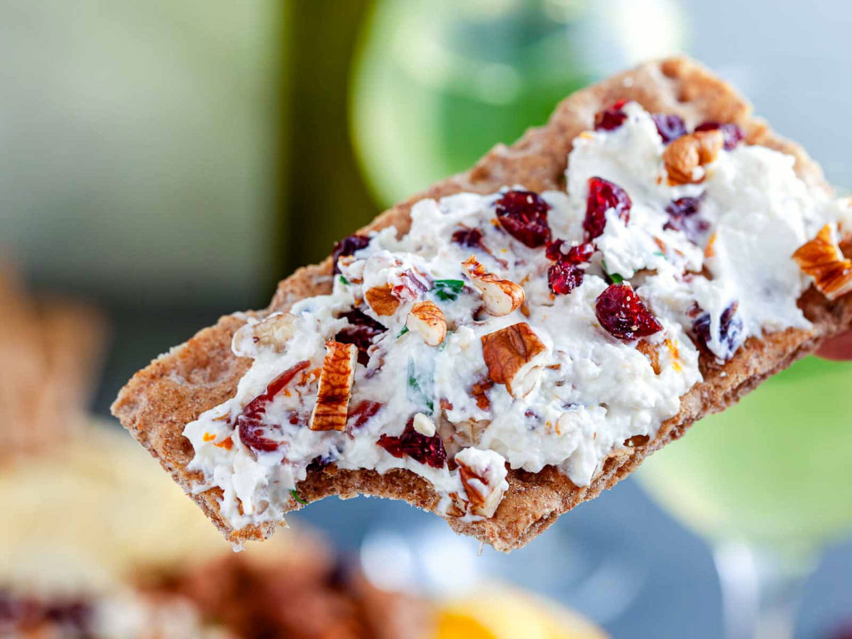 Close-up of a rye crispbread with a bite taken out, spread thickly with cream cheese, dried cranberries, and pecan pieces