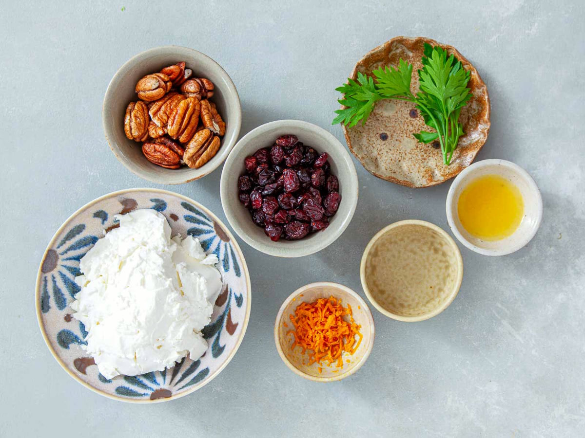 Overhead view of cream cheese, whole pecans, dried cranberries, fresh parsley, orange zest, orange juice, and water in small bowls on a gray surface.