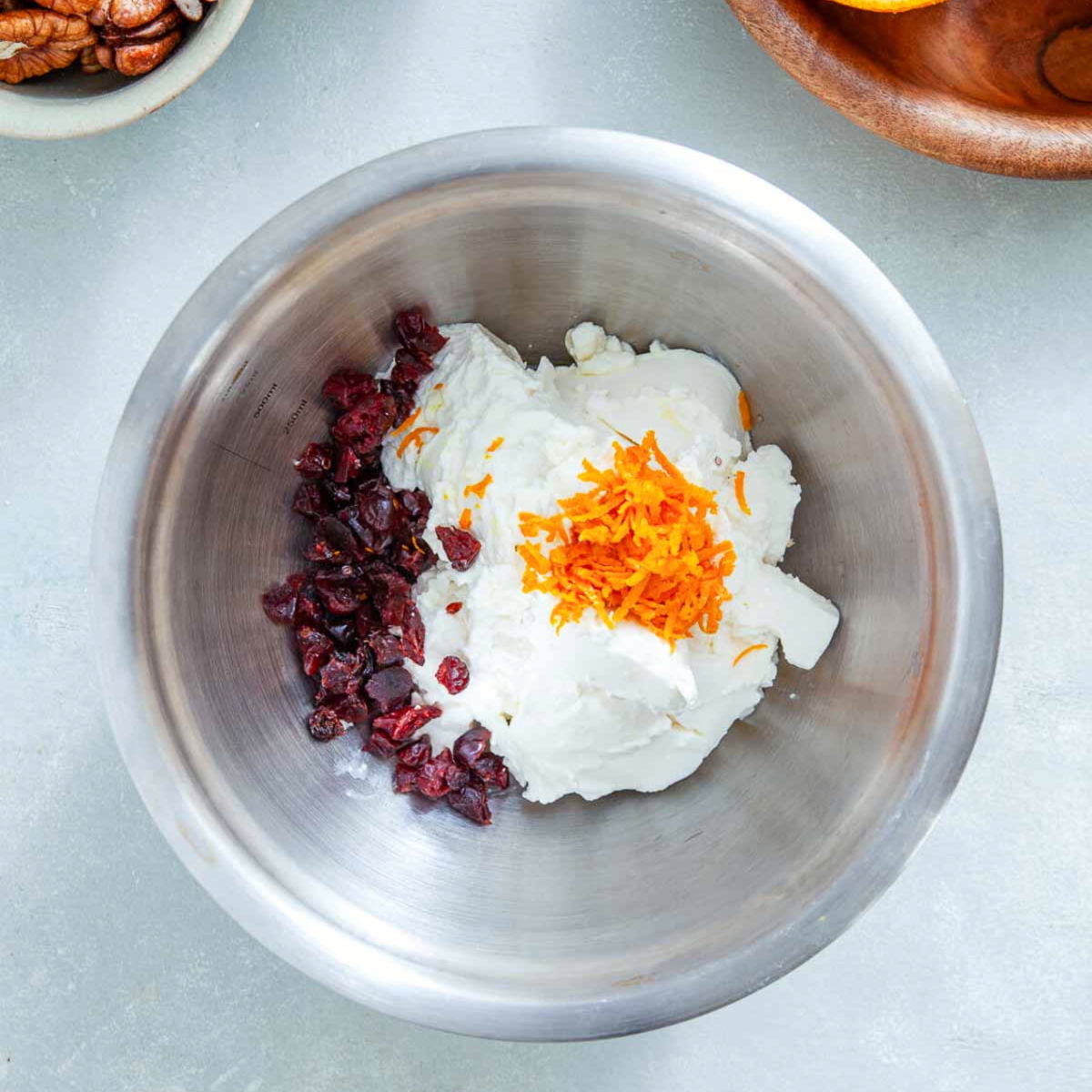  Cream cheese topped with fresh orange zest next to chopped dried cranberries in a stainless steel mixing bowl.