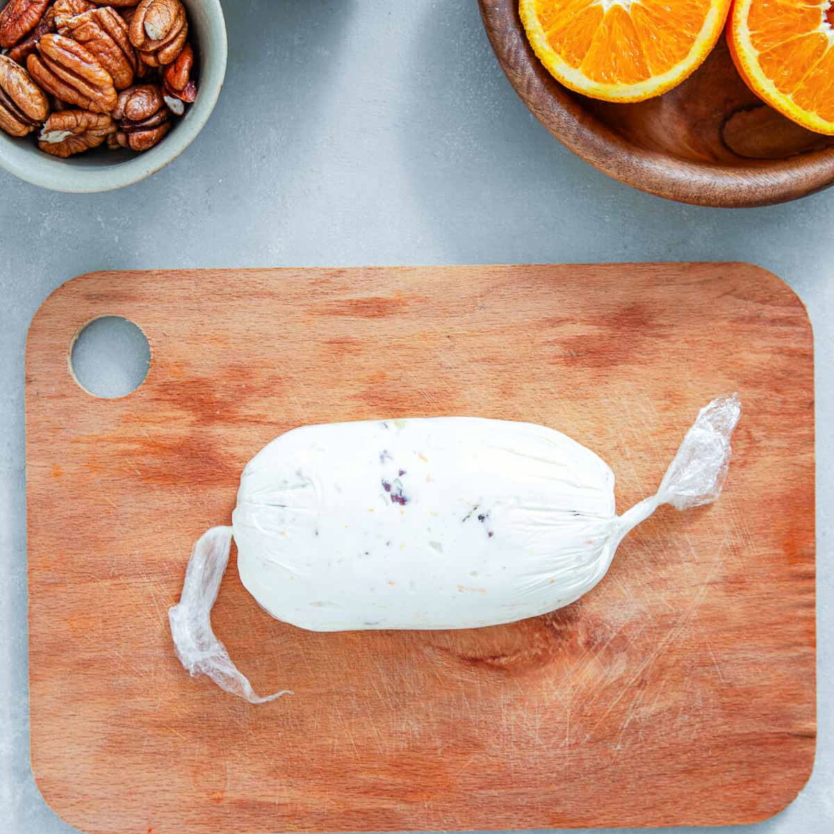 Cream cheese mixture wrapped tightly in plastic wrap and twisted at both ends, resting on a wooden cutting board beside a bowl of pecans and halved oranges.