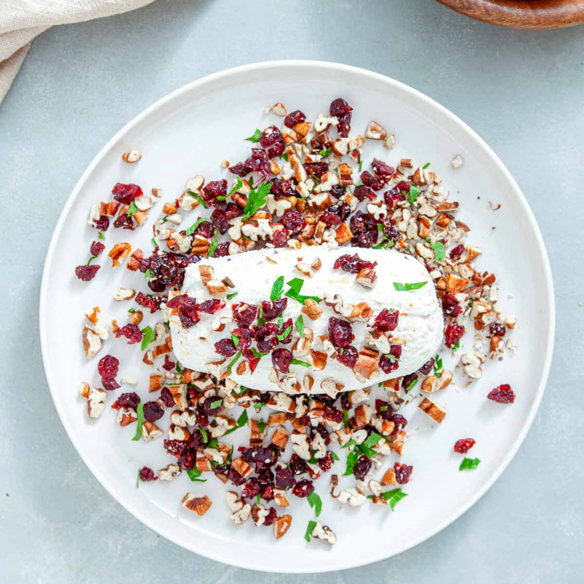 Unwrapped cream cheese log on a white plate, partly covered with chopped pecans, cranberries, and parsley, with more of the mixture scattered around it.