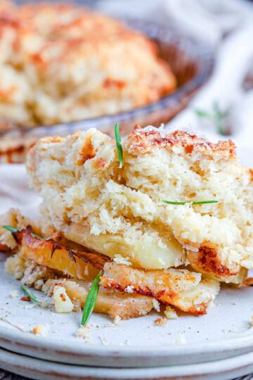 A scoop of cheesy potato cobbler on a white plate, showing the golden biscuit topping and creamy potato slices, with the baking dish blurred behind.