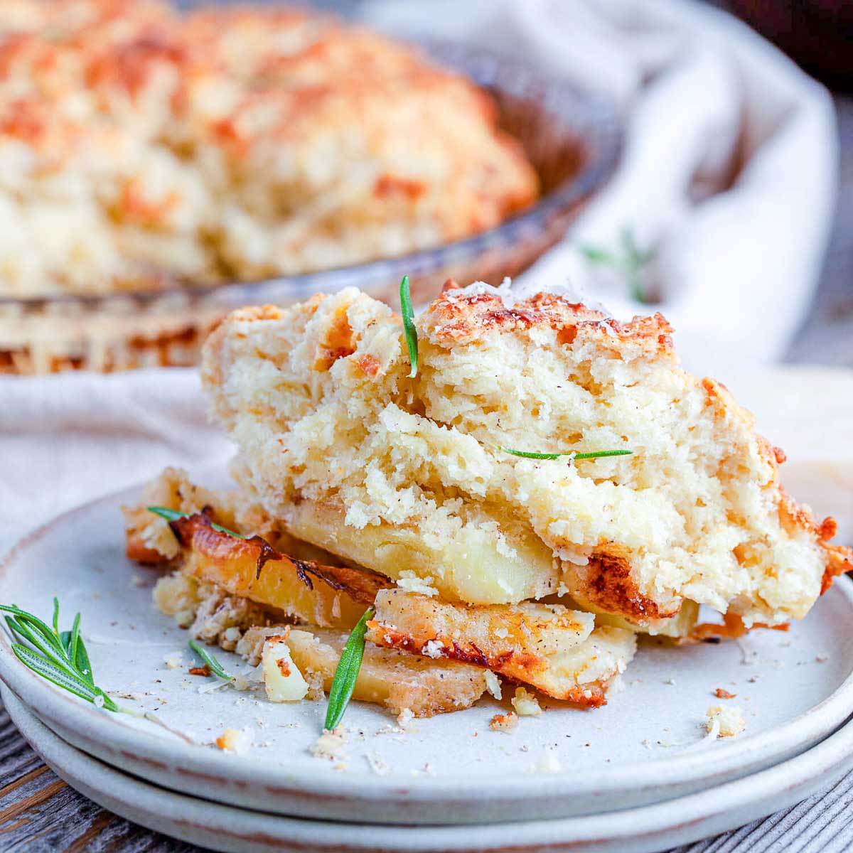 A scoop of cheesy potato cobbler on a white plate, showing the golden biscuit topping and creamy potato slices, with the baking dish blurred behind.