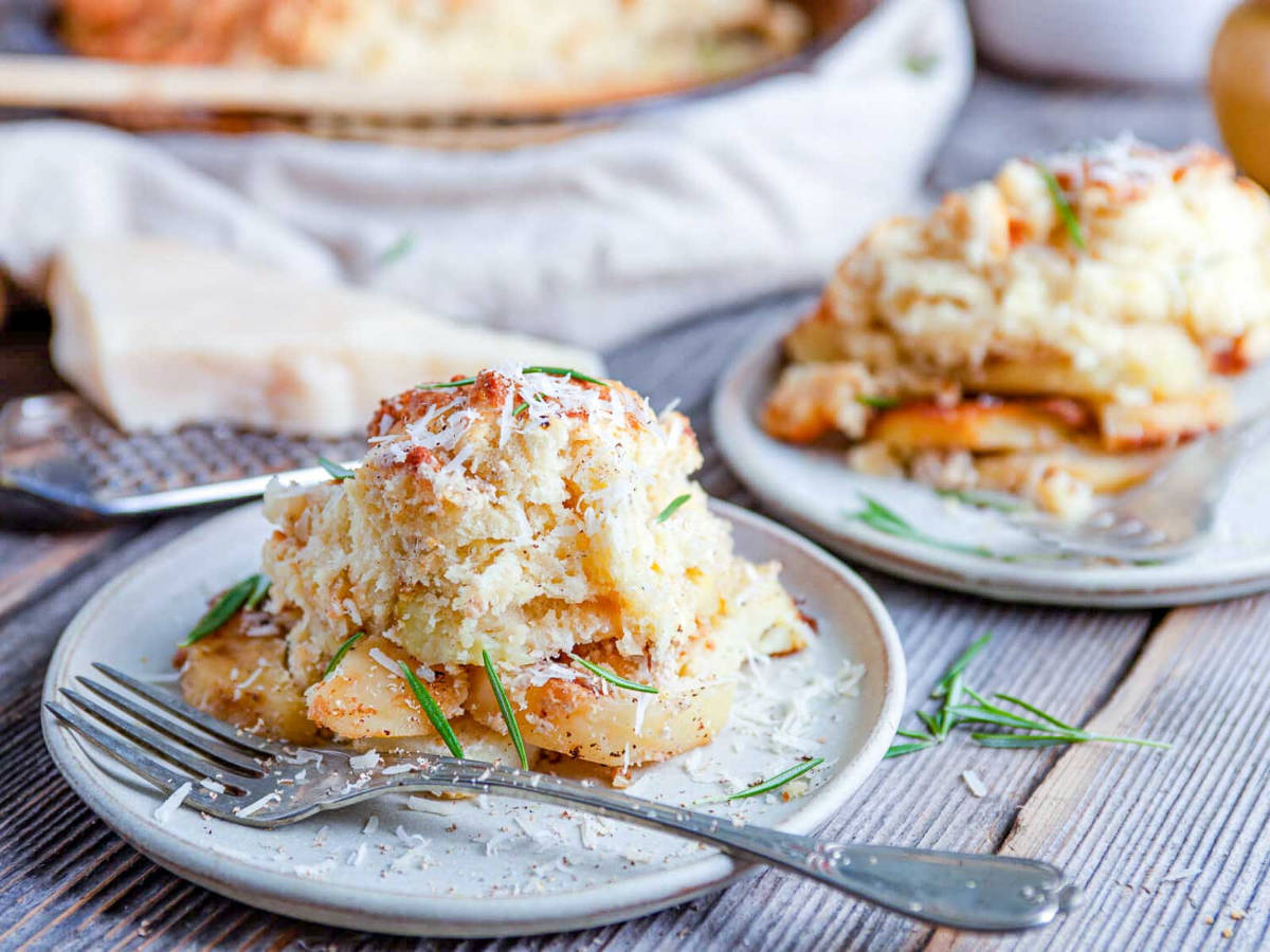 Two white plates of baked potato cobbler topped with shredded parmesan and rosemary on a weathered wood table, with a fork and a wedge of parmesan.