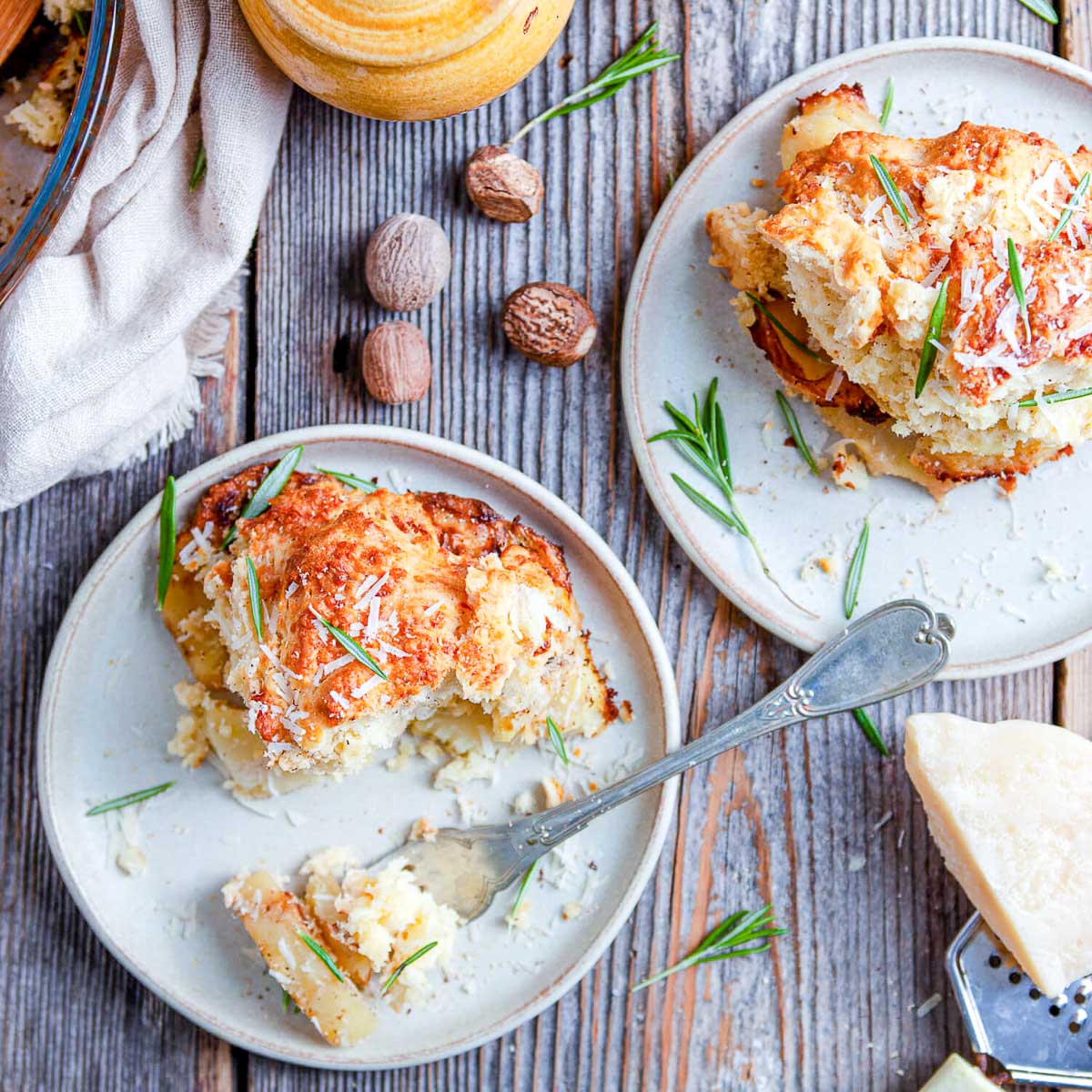 Overhead view of two plates of potato cobbler on weathered wood, with whole nutmegs, fresh rosemary, a fork, and a wedge of parmesan beside a grater.