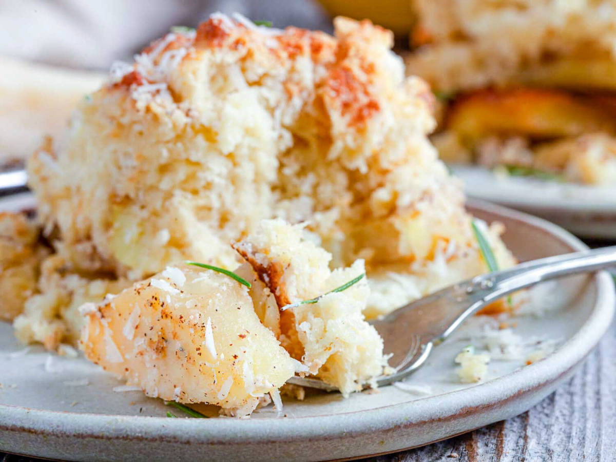 A fork lifting a bite of tender potato slices and cheesy biscuit topping from a serving of potato cobbler on a white plate.