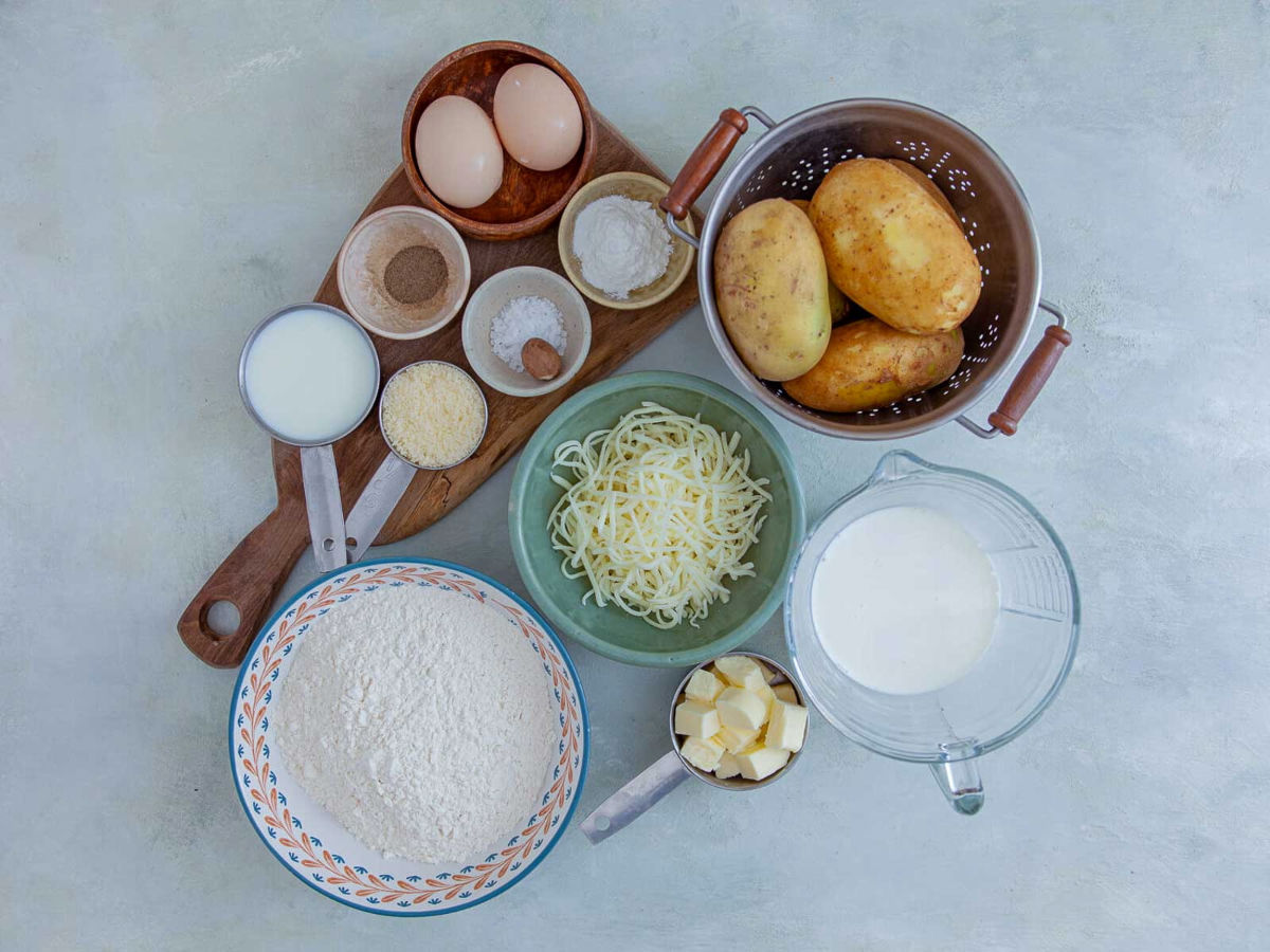 Ingredients for cheesy potato cobbler laid out on a pale surface: whole potatoes in a colander, shredded mozzarella, grated parmesan, flour, cubed butter, eggs, half and half, milk, nutmeg, and salt.