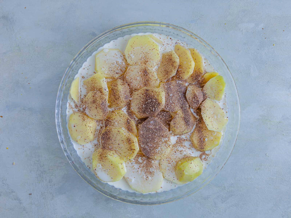 Sliced raw potatoes arranged in a glass pie plate, coated in half and half and sprinkled with ground nutmeg.